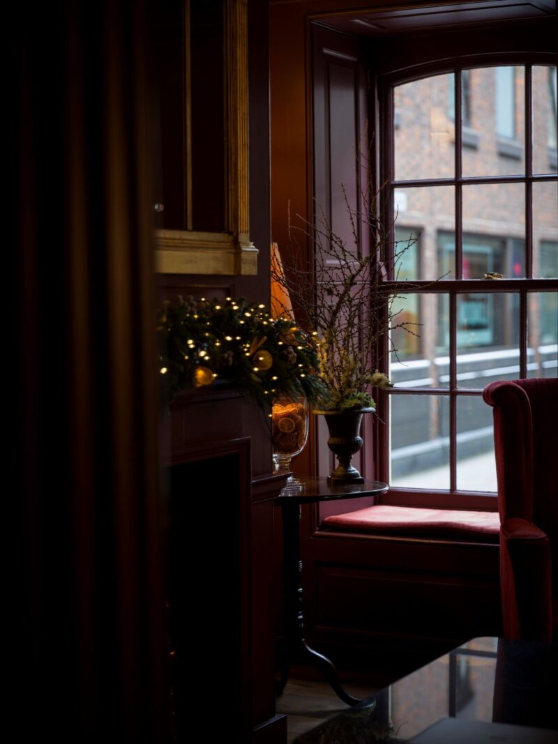 A cosy corner at Batty Langley’s with festive garland lights, a vase of winter branches, and a window seat framed by deep red wood panelling.