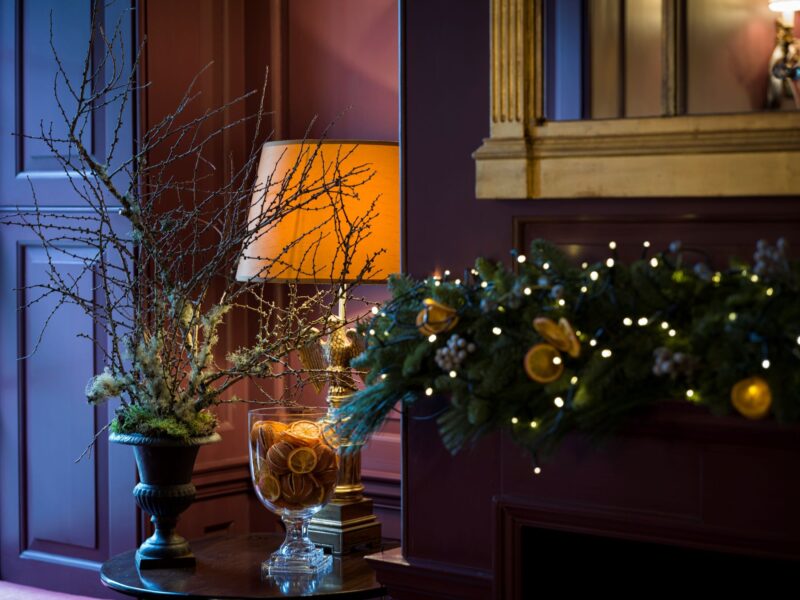 A Christmas garland with lights and dried oranges decorates a mantel at Batty Langley’s, beside a lamp, winter branches, and a glass vessel of dried citrus.