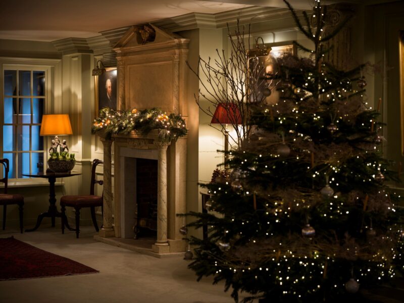 A decorated Christmas tree and a garland-lit marble fireplace inside the reception at Batty Langley’s, with warm lamps and classical portraits in the background.