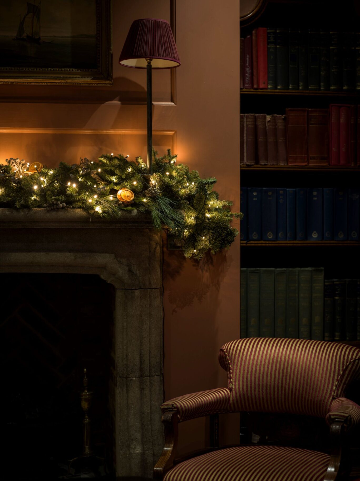 Garland-decorated fireplace with warm lights beside a striped armchair in Batty Langley’s library.