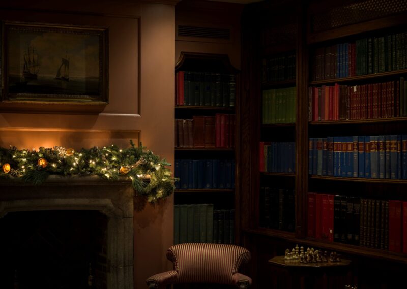 Festive garland glowing above a stone fireplace in a wood-panelled library with shelves of colourful vintage books at Batty Langley’s.