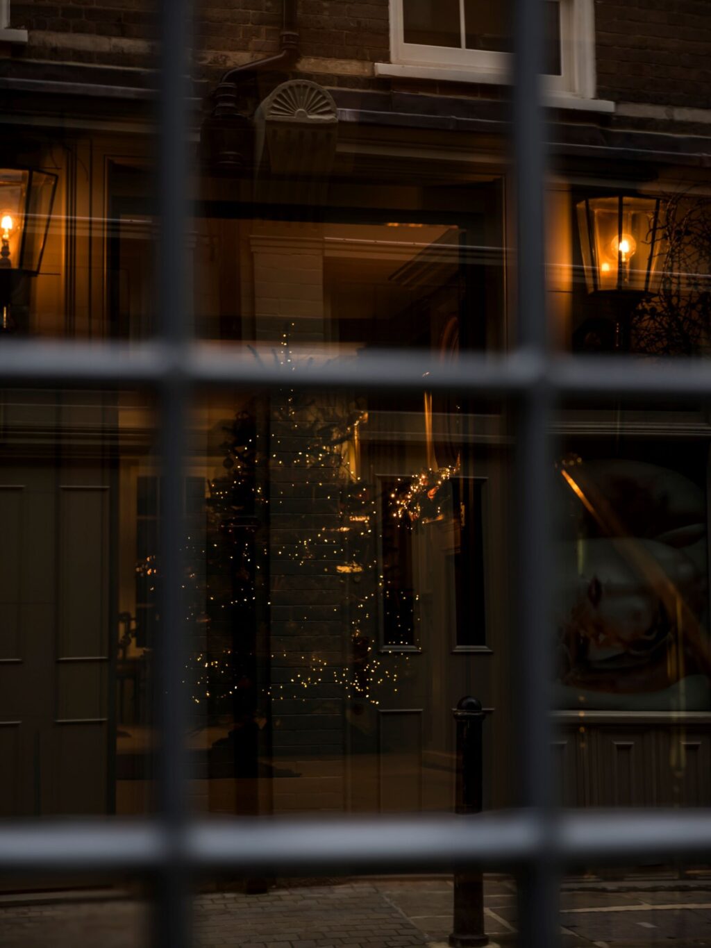 View through a paned window into Batty Langley’s, showing warm lamps and a softly lit Christmas tree reflected in the glass.
