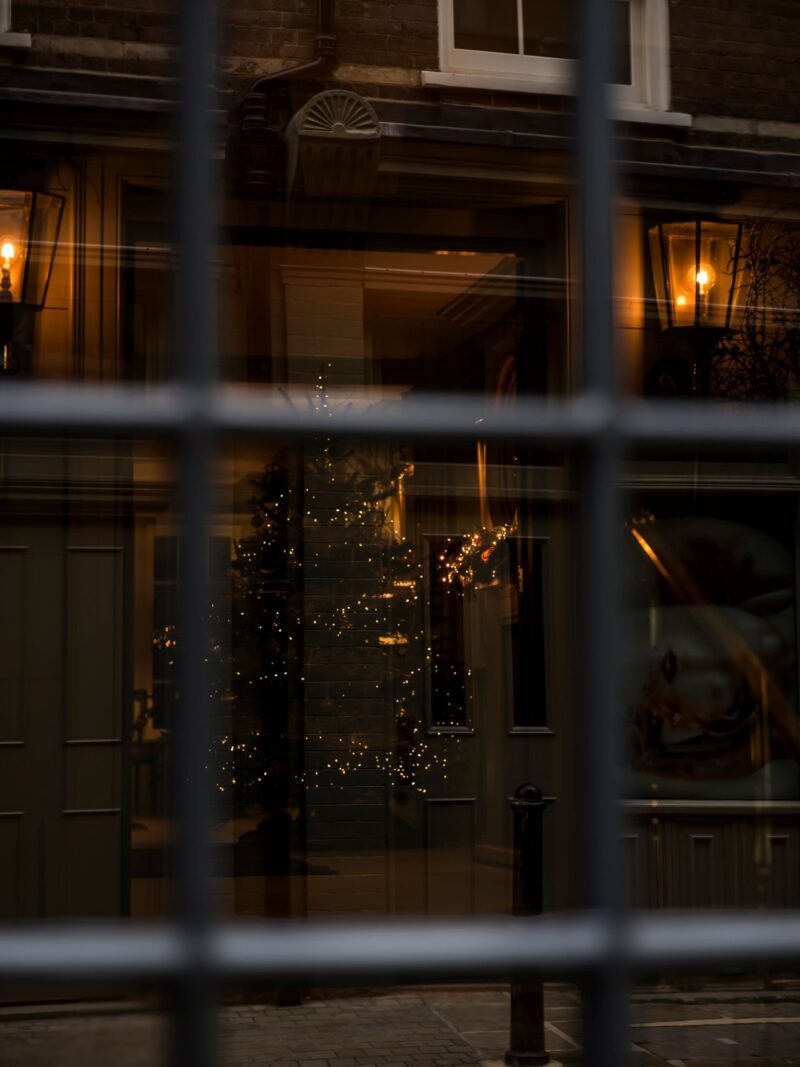 View through a paned window into Batty Langley’s, showing warm lamps and a softly lit Christmas tree reflected in the glass.
